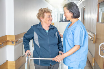 Asian female doctor reassuring mature elderly woman with walker. Two women smiling happy in the hospital aisle