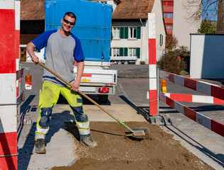 Laughing construction worker with broom standing on sand in front of vehicle and trailer looking into camera. Construction worker is happy finishing construction site of pipeline.