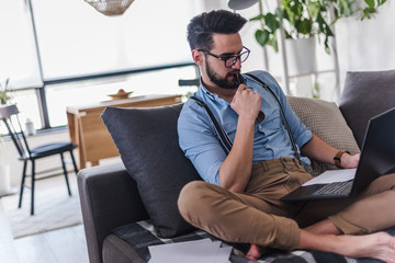 Young bearded businessman is sitting on sofa working with laptop computer. Freelancer, entrepreneur works at home.