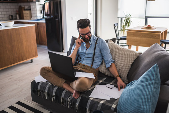 Young bearded businessman is sitting on sofa working with laptop computer. Freelancer, entrepreneur works at home.