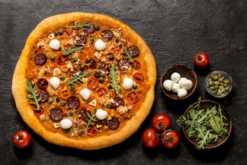 pizza with mozzarella, capers, arugula and tomatoes on black concrete background