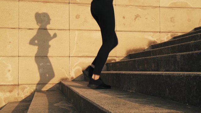 Young Woman In A Black Tracksuit Running Up The Steps. Girl On The Run. The Weather Is Sunny And Warm. A Young Girl Runs Up The Stairs During A Running Workout.