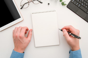 Top view man's hands with notepad on office desk. Workplace with notebook, tablet, glasses, pen and pin. Flat lay man taking notes at workplace
