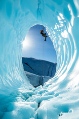 Ice climber ascending a rope over entrance of large blue ice cave on the Matanuska Glacier in...