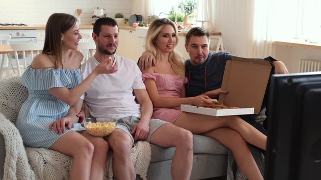 Group of friends watching TV together and eating pizza at home on sofa