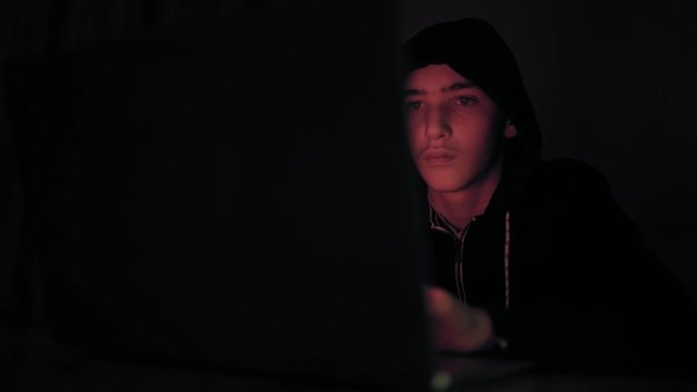 Hacker's hacking into the computer network. A young guy in a dark room typing on a laptop.  Hacker with a laptop. E-the robber. Lonely teen chatting online.
