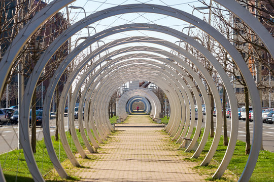 Pasillo interior formado por arcos metalicos circulares y tirantes en un paseo urbano por el que camina una persona joven con mochila 