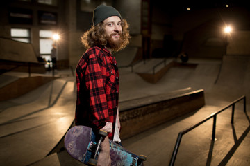 Waist up portrait of long haired man holding skateboard looking at camera standing in extreme sports park, shot with flash