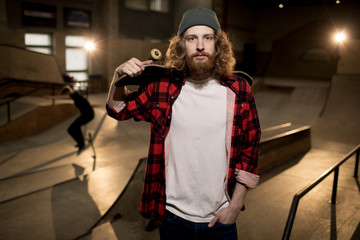 Waist up portrait of bearded man holding skateboard looking at camera standing in extreme park, shot with flash © Seventyfour