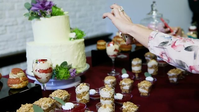 woman take pictures on phone of white cake decorated with flowers at candy bar table with assorted creamy desserts