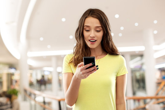 technology and people concept - smiling young woman or teenage girl in blank yellow t-shirt using smartphone over shopping mall background