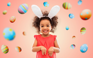 Obraz na płótnie Canvas childhood, party props and easter concept - happy little african american girl wearing bunny ears headband over colored eggs on living coral background