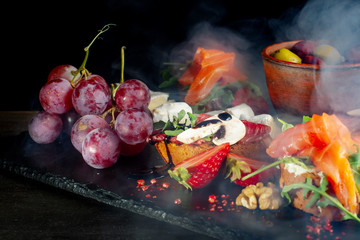 Snacks laid out on black slate, cheese, grapes, sandwiches, strawberries, nuts.