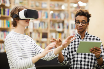 Serious handsome young African-American IT engineer using digital tablet while demonstrating 3D model to student girl in virtual reality goggles