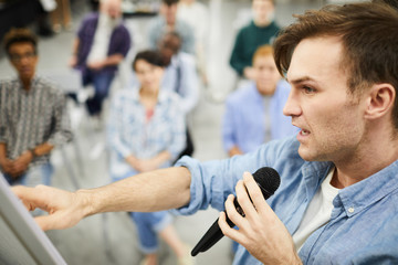 Confident handsome young man in casual shirt pointing at flip chart board and speaking into microphone while telling about powerful sales tools at business course