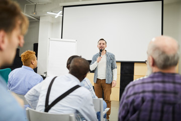 Obraz premium Content confident middle-aged startup entrepreneur with stubble standing in convention center and speaking into microphone while presenting his project at conference