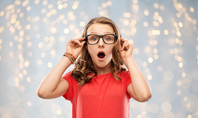 emotion, expression and people concept - surprised or shocked teenage girl with open mouth in glasses over festive lights background