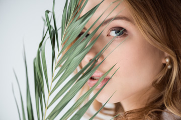 Naklejka premium pretty woman looking at camera through palm leaf on white