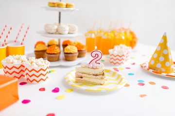 anniversary, celebration and festive concept - piece of cake with candle in shape of number two on plate at birthday party