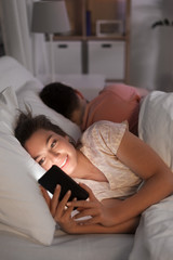 Obraz na płótnie Canvas technology, internet addiction and cheat concept - happy smiling woman using smartphone at night while her boyfriend is sleeping