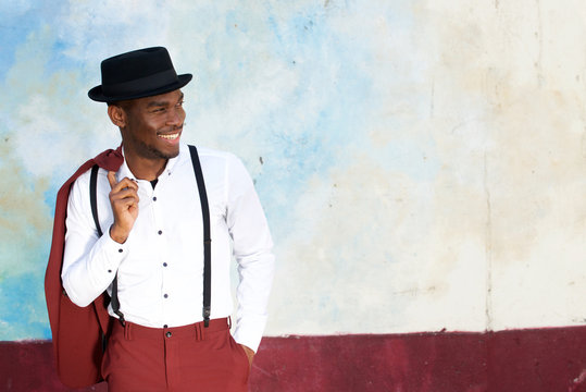 cool african american male fashion model smiling with vintage suit, suspenders and hat by wall