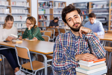 Tired man sitting with pile of books in university library on background with working fellow students