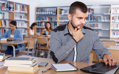 Young concentrated male student working with laptop and books in library