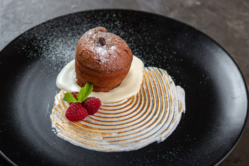 Chocolate fondant cake with vanilla cream, raspberry and mint on a black plate. Sweet muffins for...