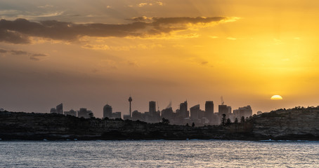 Fototapeta na wymiar Sydney, Australia - February 12, 2019: Sunset over city skyline seen from Tasman Sea. Shoreline rocky cliffs. Yellow brown sky, sun rays. 5 of 5.