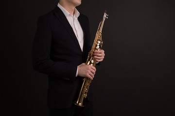 Male saxophonist in black classic suit holding a soprano saxophone standing on a black background side view - obrazy, fototapety, plakaty