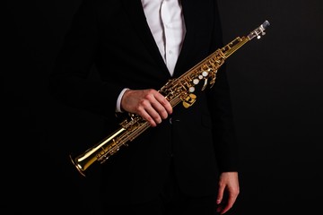 Male saxophonist in black classic suit holding a soprano saxophone standing on a black background...