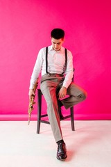 Saxophonist holding a soprano saxophone sitting on a chair on a pink background. Vertical...
