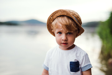 A small toddler boy standing outdoors by a river in summer, looking at camera.