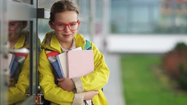 Sweet sad ten year old girl looks into the camera and then smiles. A schoolgirl with textbooks in glasses and a yellow jacket is standing near the school. Back to school