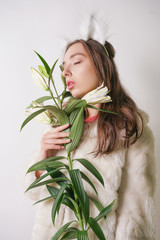 Naklejka premium cute young teen girl standing in a winter warm fur coat and cat furry ears on her head, she holds a branch of fresh lilies. woman in winter clothes with a flower on a white background in the Studio.