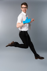 smiling businessman in glasses with paper cut thumb up jumping on grey background
