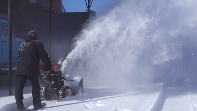 A man cleans the snow with a snowblower in sunny winter weather.
