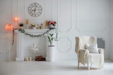 Modern white interior concept: white room with ornamental fireplace, mantelpiece, plants, large clock on the wall and white armchair