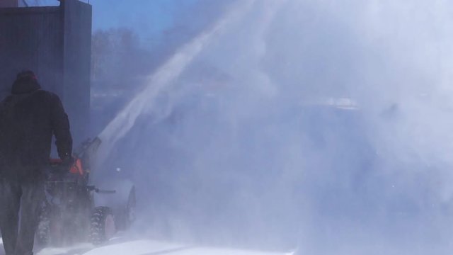 A man cleans the snow with a snowblower in sunny winter weather.