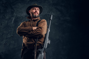 Portrait of a bearded hunter with rifle posing with his arms crossed. Studio photo against a dark wall background