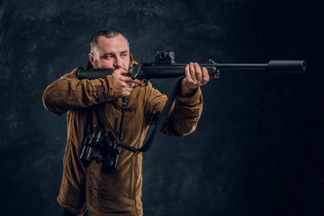 Hunter holding a rifle and aiming at his target or prey. Studio photo against a dark wall background