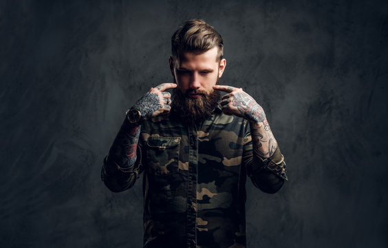 Portrait of a stylish bearded guy with tattooed hands in the military shirt. Studio photo against dark wall