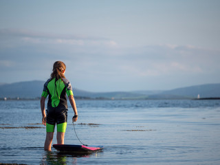 Girl in wet suit standing in the water, holding her mini surf board by a safety cord, looking into the ocean.