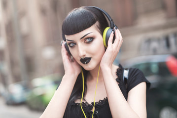 Urban goth girl listening to her favourite music over her big headphones, street in a city...