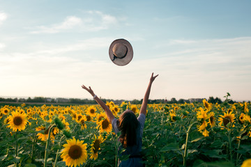 Back view of pretty young farmer girl with black hair,enjoying big crop, standing with hands up, threw straw hat in air in field of yellow sunflowers. The concept of freedom and enjoying nature.