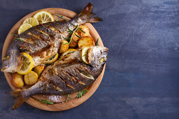 Grilled fish with roasted potatoes, lemon and rosemary on wooden tray. View from above, top, room for text