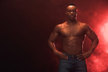 Fototapeta na wymiar sexy muscular african american man in denim looking at camera on dark with red light and copy space