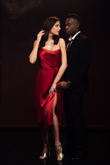 beautiful interracial couple in formal wear looking at each other and posing isolated on black