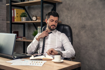 young stylish handsome businessman working at his desk in the office fixing his tie and looking at the watch