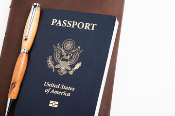 The iconic dark blue cover of an American passport with brown leather-covered journal and a ballpoint pen held together with an elastic band artistically set on a plain white background.